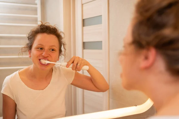 A girl bruhes teeth with white elctric toothbrush in bathroom, looking in the mirror