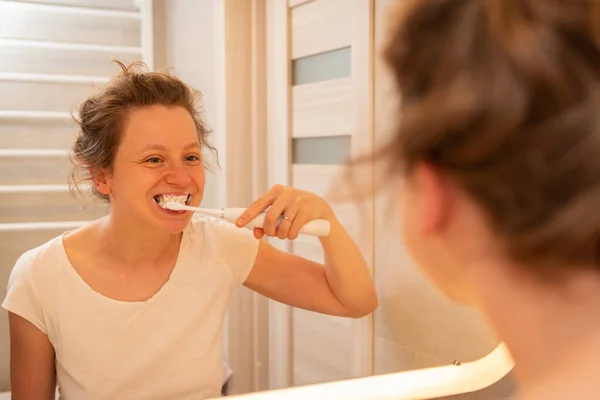 A girl bruhes teeth with white elctric toothbrush in bathroom, looking in the mirror