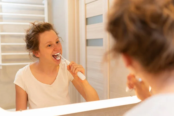 A girl bruhes teeth with white elctric toothbrush in bathroom, looking in the mirror