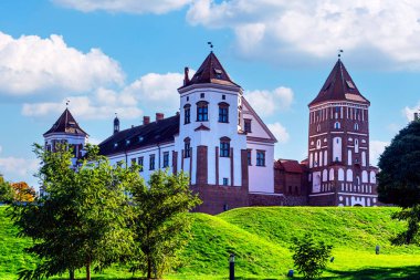 Beyaz Rusya 'daki UNESCO sahasında mavi bulutlu gökyüzünün altında tuğla kuleler ve yeşil tepe ile MIR Kalesi kompleksinin panoramik manzarası.