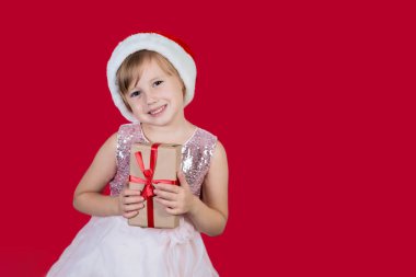 Noel Baba şapkası takmış, kameraya mutlu bir şekilde bakıp kırmızı arka planda izole edilmiş bir hediyeyi kucaklayan komik, kaktüslü, sevimli kız çocuğu. Mutlu Noel hediyeleri alışverişi.