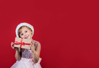 .Noel Baba şapkası takmış, elinde hediye kutusuyla kırmızı arka planda izole edilmiş mutlu bir 2021 yılını kutlayan neşeli, kaküslü çocuk. Mutlu Noel hediyeleri alışverişi.