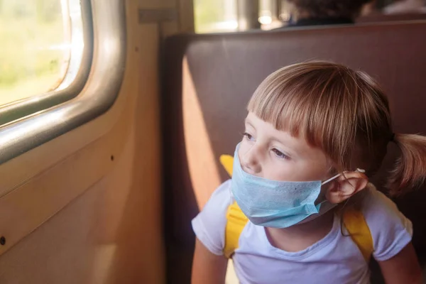 Trende beyaz tişörtlü, kot pantolonlu ve sağlık maskeli küçük bir kız oturuyor. COVID-19 'un yayılmasını önlemek için sosyal mesafeyi koruyun. İyi yolculuklar..