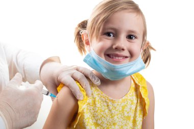 Koronavirüs çağında aşı kavramı. Doktor, beyaz arka planda sarı elbise ve yüz koruyucu maske takan sevimli gülümseyen küçük kızı aşılıyor..