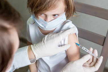 Tıbbi maskeli ve eldivenli bir doktor Coronavirus enfeksiyonuna karşı aşı yapar. Tıbbi maskeli küçük beyaz bir kızın omzuna. Kız doktora bakar.