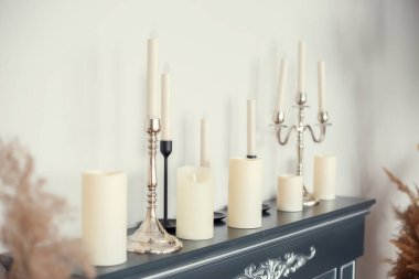 Duvarın yanındaki şömine tezgahında şamdanlı ve şamdansız çeşitli tip ve boyutlarda mumlar var.