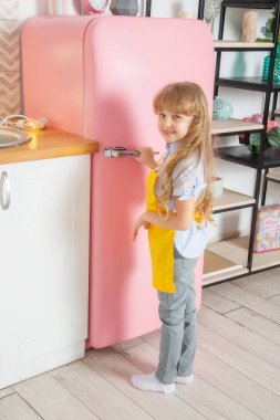 Kafkas görünümlü küçük bir kız, 7 yaşında, açık İskandinav mutfağında pembe bir buzdolabının kapısını açıyor.