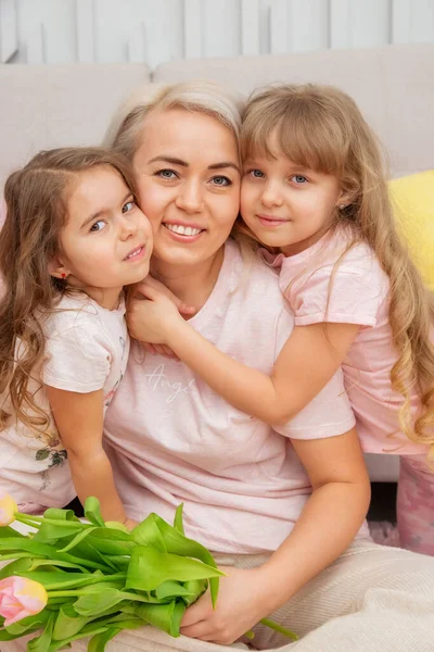 Little girls of Caucasian appearance are hugging with their mother, sitting on the floor in front of the sofa in a bright living room in a scandinavian style