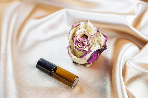 Kurumuş gül çiçekleriyle çevrili ipek kumaş kıvrımlarında koyu camdan yapılmış kozmetik ürünler depolamak için çeşitli kaplar bulunur. Parfüm, serum ya da losyon şişesi.