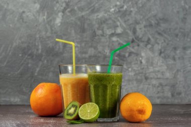 İki uzun bardak portakal suyu, muz-portakallı kivi ve gri beton arka planda meyve yarımlarıyla çevrili ıspanaklı içecek. Kilo verme ve düzgün beslenme kavramı.