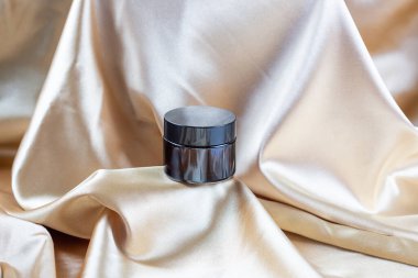 İpek kumaşın kıvrımlarında duran koyu camdan yapılmış kozmetik ürünleri depolamak için bir kap. Parfüm, serum ya da losyon şişesi.