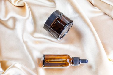 İpek kumaşın kıvrımlarında duran koyu camdan yapılmış kozmetik ürünleri depolamak için bir kap. Parfüm, serum ya da losyon şişesi.