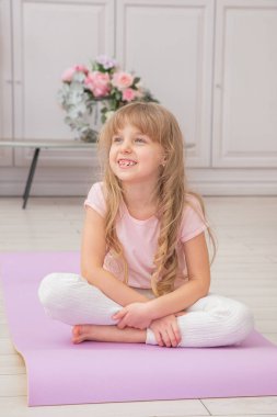 Yoga minderinde gülümseyen sevimli, oynak bir anaokulu kızı lotus pozisyonunda oturuyor. Evde yoga yapan mutlu bir çocuk..