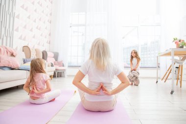 Arkadan bakınca, yoga minderinde gülümseyen sarışın anne ve sevimli küçük anaokulu kızı çeşitli egzersizler yapıyorlar. Mutlu anne küçük çocukla yoga yapıyor..