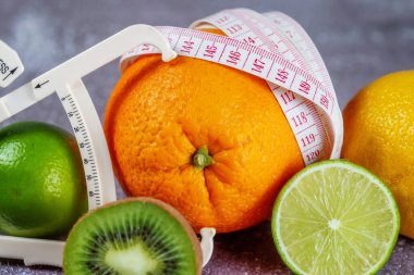 Ölçüm bandıyla sarılmış bir portakal ve gri beton arka planda taze meyvelerle çevrili bir kalibre. Zayıflatma konsepti, selüliti kaldır, şekle sok.