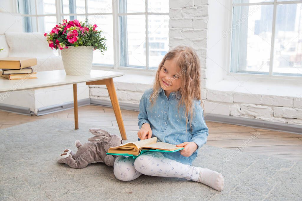 Little caucasian girl holding a book and smiling while sitting on the floor in the living room