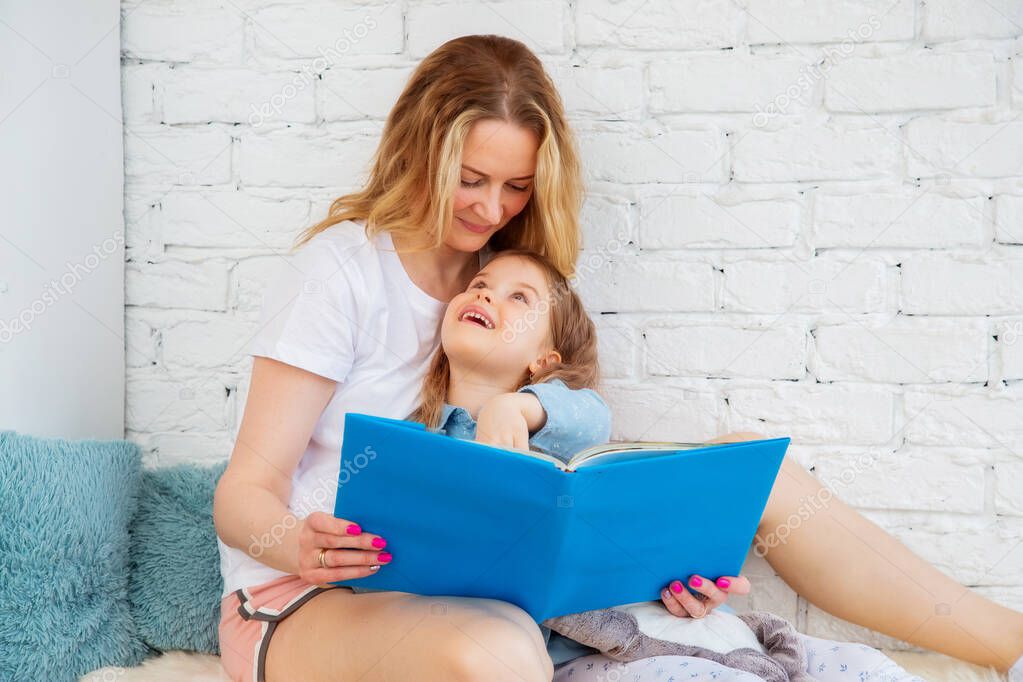 Happy Caucasian mom reads a cheerful fairy tale in a book to her daughter and smiles while sitting in the living room