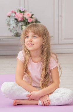 Yoga minderinde gülümseyen sevimli, oynak bir anaokulu kızı lotus pozisyonunda oturuyor. Evde yoga yapan mutlu bir çocuk..