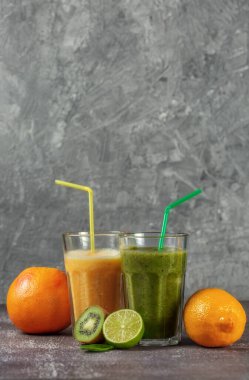 İki uzun bardak portakal suyu, muz-portakallı kivi ve gri beton arka planda meyve yarımlarıyla çevrili ıspanaklı içecek. Kilo verme ve düzgün beslenme kavramı.