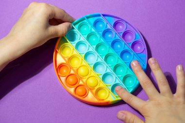 Beyaz kız duygusal anti-stres çok renkli Pop It 'i yuvarlak şekilli oyuncakla oynuyor. Çocuklar için renkli stres önleyici silikon sensörler ve mor arka planda yetişkin oyuncakları.