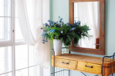 Konsol masasının üstündeki ahşap çerçevede zarif bir ayna. Pencere kenarındaki kırsal yatak odasında vazoda çiçekler var.
