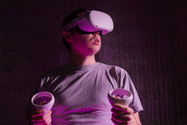 25-30 yaşlarında beyaz bir adam sanal gerçeklik kulaklığı takıyor ve neon ışıkta otururken el kol hareketi yapıyor. VR, gelecek, cihazlar, teknoloji konsepti.