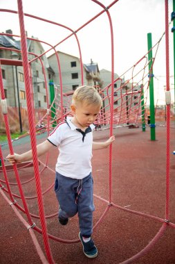 Küçük çocuk yaz aylarında evlerin arka planına karşı oyun alanında iplere tırmanıyor.