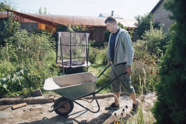 30 yaşında beyaz bir adam, yazlık evinde yolları betonla doldurmak için kum yüklemek için bir bahçe el arabası taşıyor.