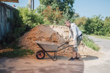 30 yaşında beyaz bir adam büyük bir kum yığınından el arabasına kum döküyor.