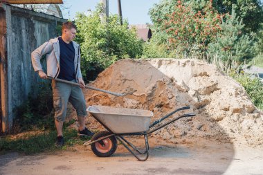 30 yaşında beyaz bir adam büyük bir kum yığınından el arabasına kum döküyor.