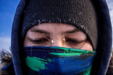 Kafkasyalı bir adamın maske gibi bir eşarbı ve kaşlarında don olan kapüşonlu kışlık ceketi yakın plan görüntüsü.