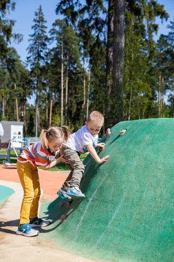 Kafkasyalı küçük kız, güneşli bir günde, çocuk parkının duvarına tırmanmak için kardeşine yardım ediyor..