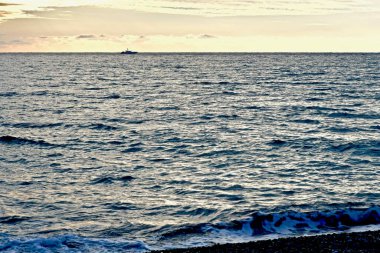 Gri denizli deniz manzaralı, bulutlu gökyüzü ve gün batımında ufukta bir gemi.