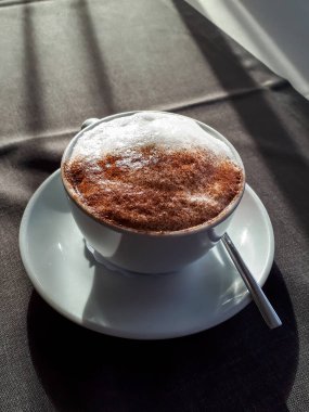 Pencerenin önündeki nötr gri masa örtüsüyle kaplı masada kapuçino kahvesi ve parlak güneş ışığı manzarası