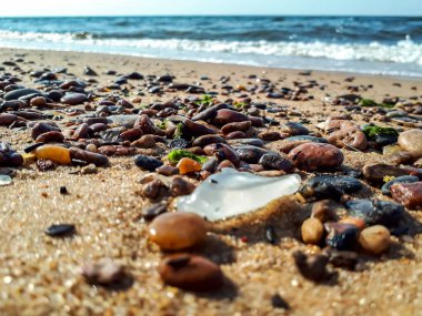 Deniz camı (plaj camı), pürüzsüz kenarları ve Baltık Denizi sahilinin kumlarında birikmiş küçük çakıl taşlarıyla görülebilir su ve arka planda dalgalar ile makro bir çekim.