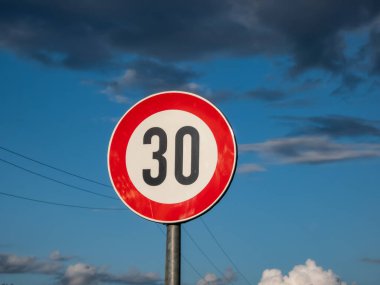Kırmızı daire içinde 30 numaralı metal yol veya trafik işareti, mavi gökyüzü ve bulutlara karşı yeni hız limitini gösterir