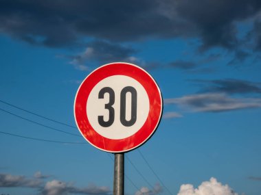 Metal road or traffic sign with number 30 in a red circle, indicating new speed limit against blue sky and clouds background clipart