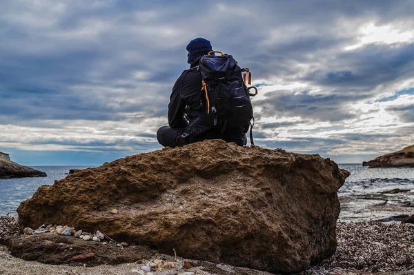 Sırt çantalı bir adam ön planda bir kayanın üzerinde oturuyor ve sana bakıyor.