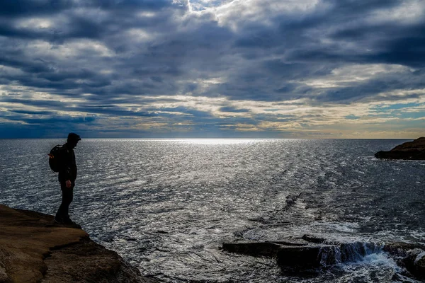 Sırt çantalı bir adam bir kayanın üzerinde durup denizi izliyor..
