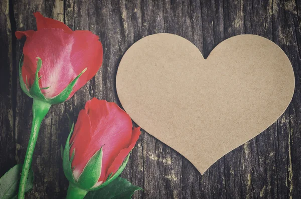 Papel marrón en blanco con forma de corazón y rosas rojas en la vieja mesa de madera — Foto de Stock