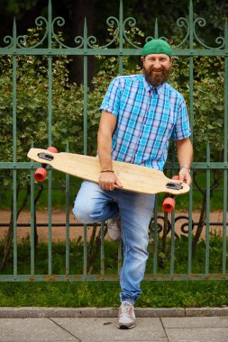 Gülümseyen hipster adam longboard ile ve moda yeşil kap üzerinde poz.