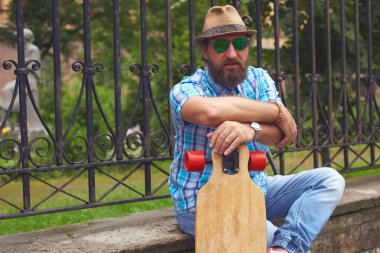 Uzun tahta ile parkta oturan hipster adam. Güneş gözlüğü ve şapka açık poz yakışıklı erkekler