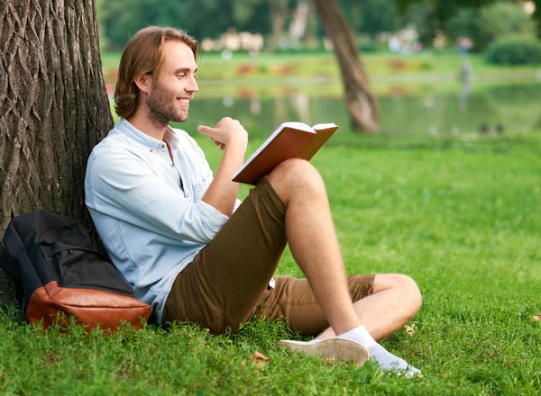 Lächelnder Student im Park des Campus liest ein Buch — Stockfoto