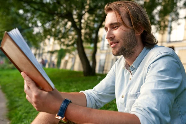 Lächelnder Strubbelmann mit einem Buch auf dem Rasen des Campus. zurück zum Schulkonzept — Stockfoto