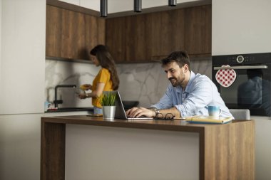 In a stylish kitchen, a man focuses on his laptop at the island while a woman prepares a meal nearby. The scene captures a casual, productive morning at home.