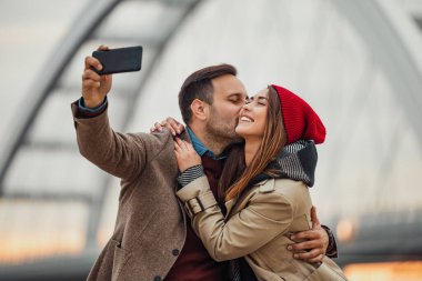 A couple embraces on a bridge while capturing a cheerful selfie. They show joy and affection, surrounded by a warm sunset glow in an urban environment.