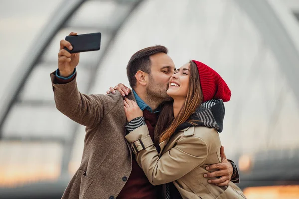 Bir çift, neşeli bir selfie çekerken köprüde kucaklaşır. Neşe ve şefkat gösterirler, kentsel bir ortamda sıcak bir günbatımı parıltısıyla çevrili..