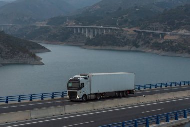 Dondurulmuş yarı römorklu bir kamyon bir su deposunun üzerinden geçiyor ve arka planda otoyol var..