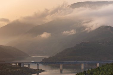 Dondurulmuş kamyonlar, gün doğumunda dağlarla ve alçak bulutlu ormanlarla çevrili bir bataklığın üzerindeki köprüde yol alıyorlar..