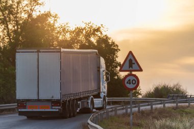 Truck with semi-trailer for general cargo reaching a limited speed crossing at sunset.